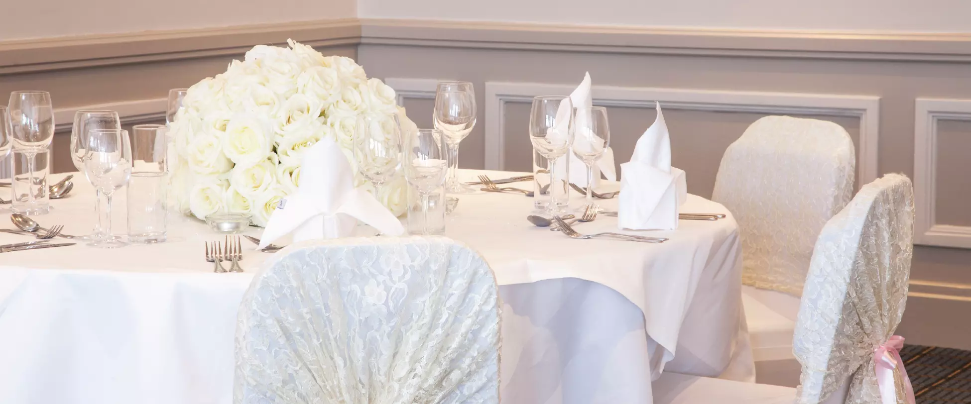 A circular table set for a wedding with white table cloth, white chair covers, white flowers and dinnerware..