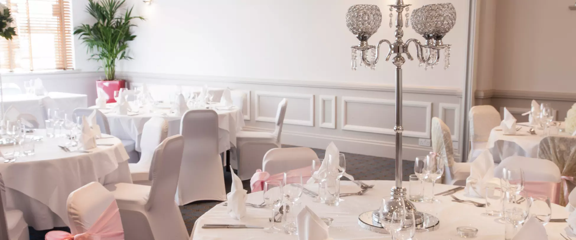 Wedding tables set up with white table cloths, pink ribbons, flowers and large silver centrepieces.